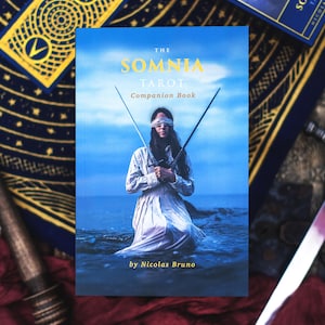 May include: The Somnia Tarot Companion Book by Nicolas Bruno. The cover shows a person with a blindfold holding two swords, set against a blue sky and ocean backdrop. Additional tarot cards are visible in the background, with gold and navy blue designs.