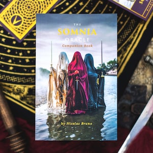 May include: The Somnia Oracle Companion Book by Nicolas Bruno. The cover shows three robed figures in water, holding swords. Tarot cards with gold and purple details surround the book, set against a patterned background.