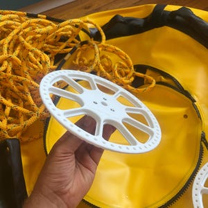 Könnte beinhalten: Eine weiße, kreisförmige Kunststoffspule mit Speichendesign und perforierten Rändern, gehalten über einer leuchtend gelben Ausrüstungstasche, die ein aufgewickeltes gelbes Seil mit schwarzen Sprenkeln enthält. Die Spule hat eine zentrale Nabe.