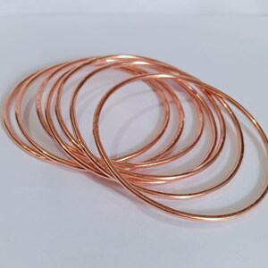 May include: A set of seven thin, circular bangles made of polished copper. Each bangle features a smooth, rounded surface with a metallic, reddish-brown finish. The bangles are arranged in an overlapping, stacked formation, creating a series of concentric circles that highlight the reflective quality of the copper material.