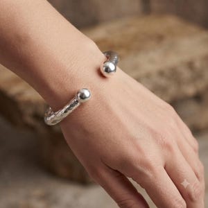 May include: A silver-toned cuff bracelet featuring a hammered, textured surface and two smooth, polished spherical ends. The open-ended design wraps around the wrist, showcasing a reflective metallic finish with a distinct contrast between the dimpled band and the rounded, shiny terminals.