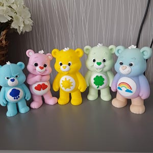 May include: Five Care Bear figurines standing in a row. From left to right: a light blue bear with a blue rain cloud, a pink bear with two red hearts, a yellow bear with a yellow sun, a light green bear with a green clover, and a pastel bear with a rainbow.