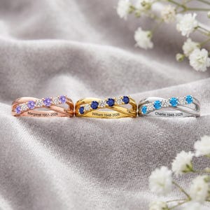 Op de afbeelding: Drie gekruiste ringen in roségoud, geelgoud en zilverkleur. Elke ring is voorzien van afwisselende ronde edelstenen en heldere stenen. De roségouden ring is gegraveerd met Margaret 1951-2026, de geelgouden met William 1948-2026 en de zilveren met Charlie 1948-2026.