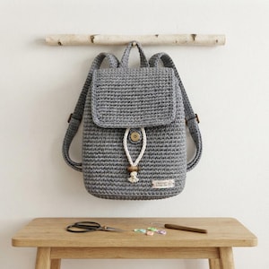 May include: A grey crocheted backpack featuring a flap closure with a white cord drawstring, a metallic snap button, and a small wooden bead accent. A rectangular fabric label with the text Crochet Mama Zen is attached to the lower right corner.