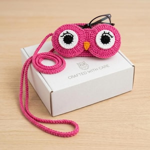 Peut inclure: Un étui à lunettes en crochet rose vif en forme de hibou avec une longue lanière assortie. L'étui présente de grands yeux blancs et noirs, un petit bec orange et des lunettes à monture noire posées dessus. Il repose sur une boîte blanche.
