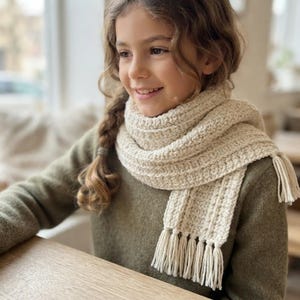 May include: A cream-colored crocheted scarf featuring a ribbed texture and finished with fringe tassels at both ends. The scarf is wrapped around the neck, showcasing the intricate yarn work and the soft, neutral tone of the handmade accessory.