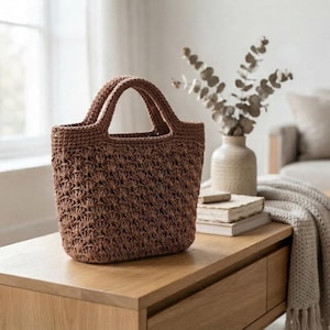 May include: A medium-brown, hand-crocheted tote bag featuring a textured shell stitch pattern throughout the body. The bag has two integrated, sturdy handles and a structured, open-top design, crafted from a durable, woven fiber material.