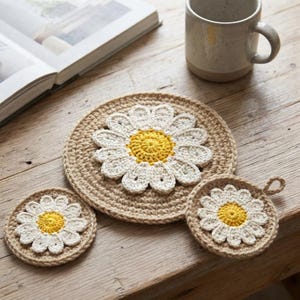 Può includere: Un set di tre sottobicchieri rotondi all'uncinetto e un tappetino con un motivo a margherita con centro giallo e petali bianchi su base color marrone chiaro. Gli articoli sono circolari con una finitura in filato testurizzato. Un occhiello.