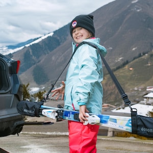 May include: A pair of skis held in a black carrying strap system with the text 6000B.C. printed on the ends. The skis are white and blue with integrated bindings, suspended by an adjustable black shoulder strap for transport and storage.