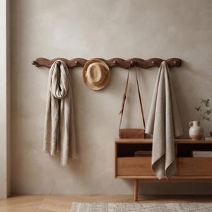 May include: A dark brown wooden wall-mounted coat rack with a wavy, scalloped edge design featuring eight rounded pegs. The rack holds a beige linen scarf, a straw hat, a brown leather crossbody bag, and a folded beige textile.