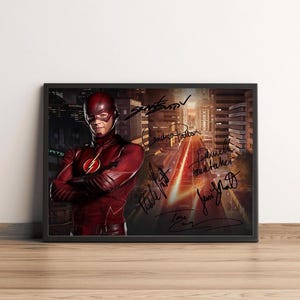 Op de afbeelding: Een ingelijste print met The Flash in een rood pak met een geel bliksemembleem, staand voor een stadsstraat bij zonsondergang. De afbeelding bevat meerdere handtekeningen in zwarte inkt die over de rechterkant van de compositie zijn geplaatst.