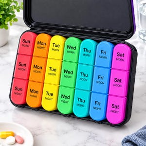 May include: A black plastic pill organizer case with a hinged lid, containing seven vertical columns of colorful compartments. Each column represents a day of the week, labeled with the day and MORN, NOON, or NIGHT in black text on the lids.