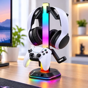 May include: A black gaming headset and controller stand featuring a vertical RGB light bar and a base with integrated LED lighting. The stand holds a white wireless gaming controller and a pair of white over-ear headphones with black ear cushions.