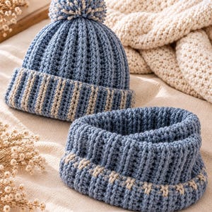 May include: A blue and cream knit beanie with a matching pom-pom and a coordinating blue and cream knit cowl neck scarf. Both items feature a vertical ribbed texture and a two-tone color pattern with a folded brim on the beanie.
