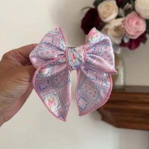 Puede incluir: Un lazo para el cabello de tela satinada con un patrón de patchwork de cuadrados rosa claro y azul con motivos florales y geométricos. El lazo tiene un borde ribeteado en rosa y un nudo central, creando una forma estructurada con dos caídas.