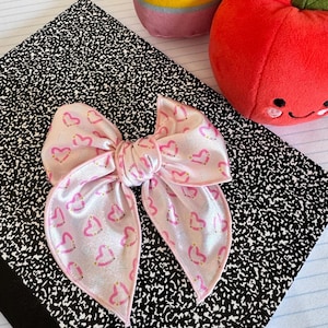 May include: A light pink satin hair bow featuring a repeating pattern of small pink hearts with yellow and white accents. The bow has a knotted center and pointed ends, with a smooth, shiny finish that reflects light across its surface.