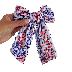 Puede incluir: Un lazo de tela blanca adornado con lentejuelas rojas, blancas y azules. El lazo está sujeto por una mano sobre un fondo blanco. Las lentejuelas están dispersas por todo el lazo, creando un diseño festivo y llamativo. Este accesorio es adecuado para varias ocasiones.