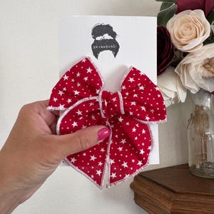 Puede incluir: Lazo para el pelo rojo con estampado de estrellas blancas, sostenido en una mano. El lazo está sujeto a una tarjeta blanca con el texto "BRYNNBANDS" y una silueta de peinado femenino. Accesorio decorativo para el cabello.