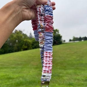Op de afbeelding: Een stoffen lanyard met een geplooid scrunchie-ontwerp, gemaakt van een patchwork van diverse textielsoorten met patronen, waaronder rood-wit gingham, bloemenprints en effen lichtblauwe secties, afgewerkt met een zilverkleurige metalen sluiting.