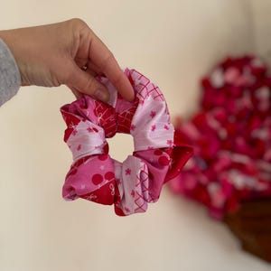 Puede incluir: Una goma para el pelo rosa y roja hecha de varias telas estampadas, incluyendo lunares, diseños florales y patrones entrecruzados. La goma se sostiene sobre un fondo neutro, mostrando sus colores y texturas vibrantes.