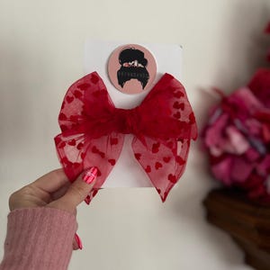 May include: A red hair bow with a heart pattern, attached to a white card. The bow is made of sheer fabric and has a central knot. A round sticker with the text "BRYNNBANDS" is also attached.