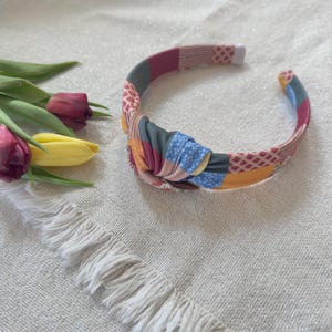 May include: A colorful fabric headband with a knotted design. The headband features a patchwork pattern in shades of pink, blue, yellow, and green. The headband is a stylish accessory for any outfit.