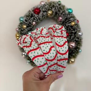 May include: Three white headbands with a red and green pattern are held in front of a Christmas wreath. The headbands have a repeating design of red lines and green speckles. The wreath is green with colorful ornaments.