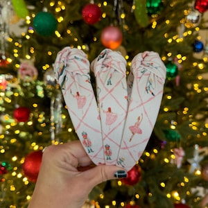 May include: Three white headbands with a pink ballet-themed pattern are held in front of a Christmas tree. The pattern includes ballerinas, nutcrackers, and a crisscross design. The headbands are knotted at the top.