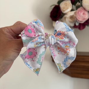 Puede incluir: Un lazo para el cabello de tela con fondo blanco y un patrón repetitivo de formas hexagonales coloridas en tonos rosa, azul, naranja y amarillo. El lazo tiene un centro anudado y bordes terminados con costuras visibles en todo el diseño.