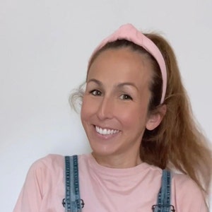May include: A woman with long brown hair is wearing a pink knotted headband and a pink t-shirt with denim overalls.