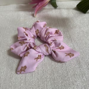 Peut inclure: Un chouchou rose clair avec un nœud, orné d'un motif répété de bonhommes en pain d'épices marron. L'accessoire pour cheveux est en tissu doux et convient à diverses coiffures. Une fleur de lys rose est en arrière-plan.