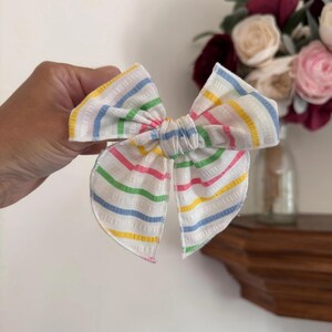 Op de afbeelding: Een stoffen haarstrik met een witte basis en dunne, horizontale strepen in geel, lichtblauw, roze en groen. De strik heeft een textuur met een licht geribbeld oppervlak en een geknoopt midden, wat zorgt voor een symmetrische vorm.