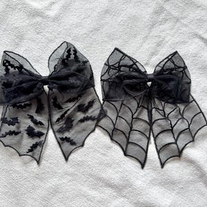 Puede incluir: Dos lazos para el pelo de tela negra transparente. Uno presenta un diseño de murciélago y el otro un patrón de telaraña. Los lazos están atados en el centro con una cinta negra. Accesorios de Halloween.