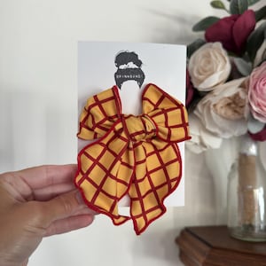 May include: A yellow fabric hair bow with a dark red grid pattern and red stitched edges. The bow is attached to a white display card featuring a black line drawing of a person with a bun hairstyle and the text BRYNNBANDS printed in black.