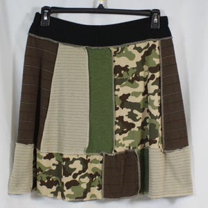 May include: A patchwork skirt with a black waistband. The skirt features panels of brown, tan, green, and camouflage fabric. The skirt is hanging on a black hanger against a white background.