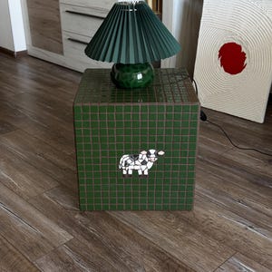 May include: A square side table covered in dark green square tiles featuring a small mosaic cow in the center. A dark green pleated lamp shade sits on a matching dark green ceramic base on top of the table, creating a cohesive lighting piece.