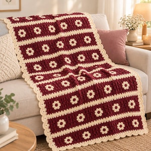 May include: A rectangular crochet blanket featuring alternating horizontal stripes of deep burgundy and cream. The cream stripes are decorated with a repeating pattern of small, six-petaled flowers. The blanket has a scalloped cream border.