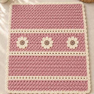 May include: A rectangular crochet blanket featuring a textured dusty rose stitch pattern, accented by two horizontal cream-colored lace-style bands and three central cream-colored flower motifs with brown centers, finished with a scalloped cream border.