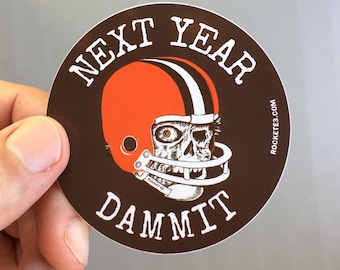 May include: A round sticker with a brown background and white text that reads "Next Year Dammit". A cartoon skull wearing a brown and white football helmet is in the center of the sticker. The text "ROCKETEB3.COM" is printed in white on the right side of the sticker.