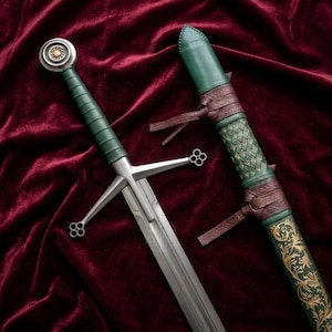 May include: A steel sword with a green leather-wrapped grip, a circular pommel with a gold center, and a crossguard with four-leaf clover terminals. Beside it lies a matching green leather scabbard featuring brown leather straps and gold filigree.