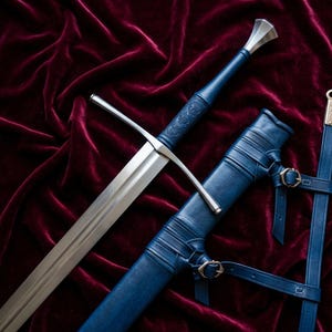 May include: A longsword with a polished steel blade, a straight crossguard, and a blue leather-wrapped hilt featuring embossed detailing. The sword is accompanied by a matching blue leather scabbard with integrated straps and metal buckles.