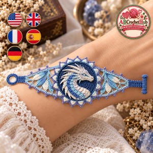 Op de afbeelding: Een blauw en wit gehaakte armband met een centraal rond medaillon met een gedetailleerd drakenmotief. De band heeft een ingewikkeld kantachtig patroon met een lus aan het ene uiteinde en een T-sluiting aan het andere uiteinde.