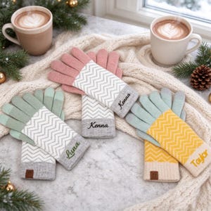 Könnte beinhalten: Drei Paar Strickhandschuhe mit Zickzackmuster auf den Handflächen und einfarbigen Fingern. Ein Paar ist rosa mit dem Namen Kenna, eines hellgrün mit dem Namen Luna und eines gelb mit dem Namen Taylor, der auf die Bündchen gestickt ist.