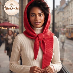 May include: A bright red knitted hooded scarf with a textured stitch pattern, tied in a knot at the front, worn over a cream-coloured ribbed turtleneck jumper. The image includes a circular logo in the top left corner with the text AshoCrochet.