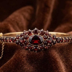 Puede incluir: Un brazalete rígido articulado en tono dorado con una piedra granate central de color rojo intenso en talla ovalada, rodeada por un grupo de granates más pequeños en talla redonda. La banda también tiene una hilera de granates redondos.