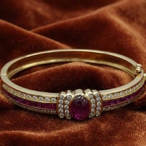May include: A gold hinged bangle bracelet featuring a central oval-cut deep red gemstone surrounded by small clear stones. The band is accented with a row of channel-set square red stones flanked by two rows of small clear round stones.