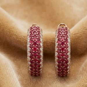 May include: A pair of hoop earrings crafted from rose gold, featuring a central band of round, deep red rubies flanked by two outer rows of small, clear white diamonds, creating a pavé-set design with a polished, metallic finish.