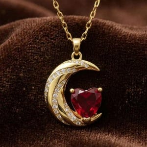 May include: A gold necklace featuring a crescent moon pendant encrusted with small clear stones. A deep red heart-shaped gemstone is set within the curve of the moon. The pendant hangs from a delicate gold chain, creating a refined jewelry piece.