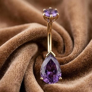 Pode incluir: Um piercing de umbigo em barra curva de tom dourado com uma pedra preciosa roxa redonda no topo e uma pedra roxa maior em forma de gota na base, ambas presas por garras de metal numa haste de metal polido de cor dourada.