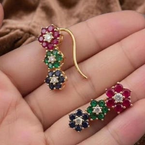 May include: A pair of gold-tone earrings featuring three flower-shaped clusters of gemstones. Each cluster contains six outer stones surrounding a single clear center stone. The colors include red, green, and blue stones arranged in a vertical line.